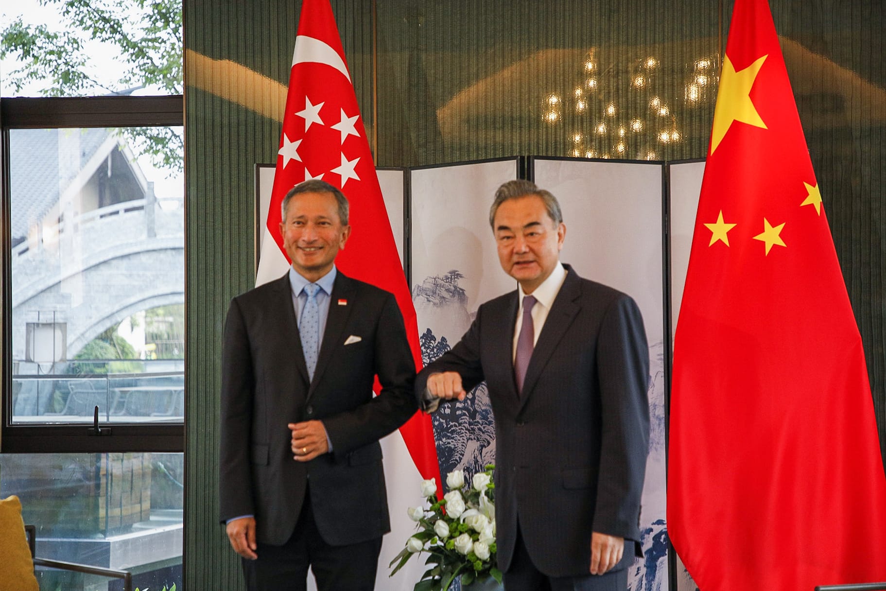 Two men in suits bump elbows near Singapore and China flags.